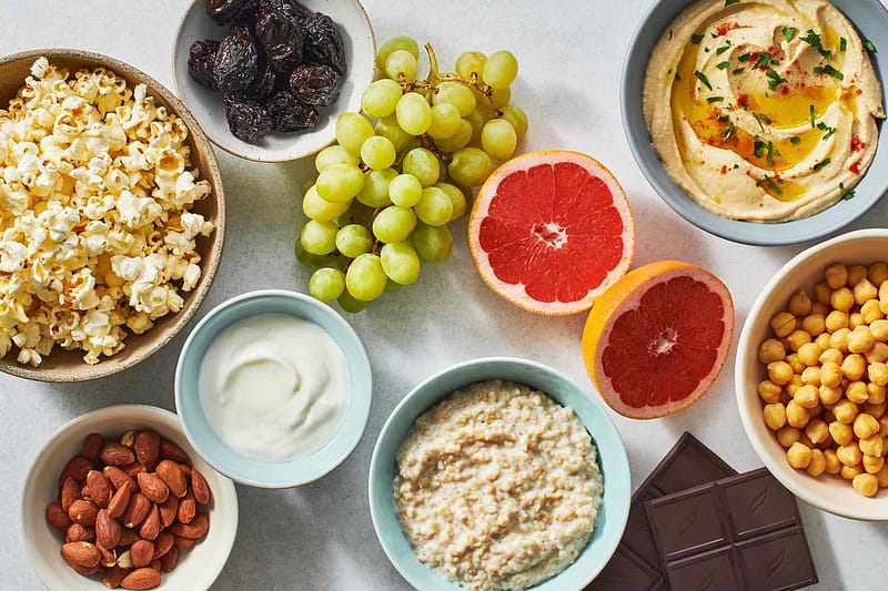 Healthy snacks including popcorn, yogurt, almonds, grapes, grapefruit, hummus, chickpeas, and oatmeal arranged on a table.